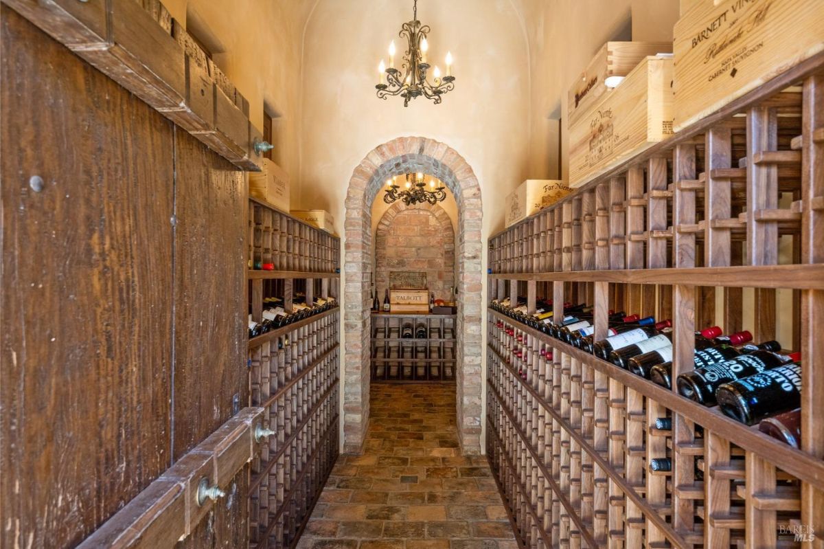 Wine cellar with brick flooring, arched brick doorways, and custom wooden wine racks lining both walls. Features a chandelier providing ambient lighting and crates of wine stored on top of the racks.