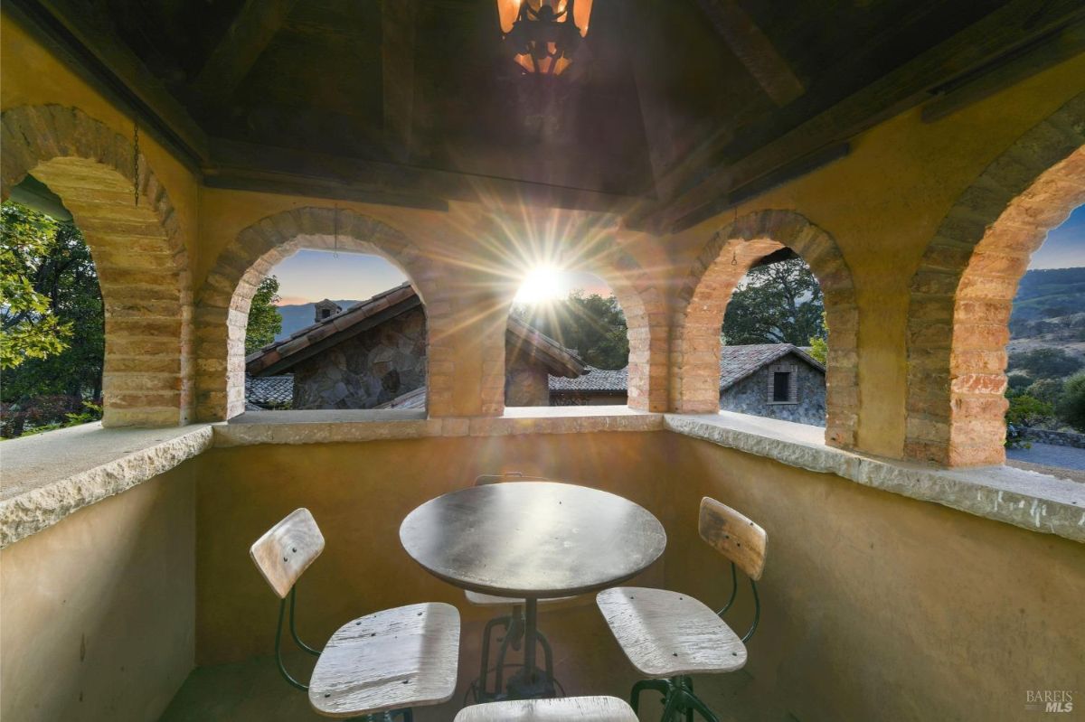 Outdoor nook with arched brick openings, a small round table, and chairs, capturing a scenic view with sunlight streaming through, surrounded by rustic structures.