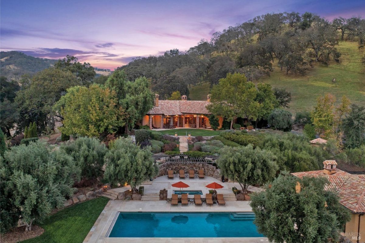 Expansive estate featuring a rectangular pool with adjacent lounge areas, surrounded by lush landscaping and trees. The main residence, with a tiled roof and arched doorways, is nestled into a hillside and overlooks the pool and garden spaces at sunset.