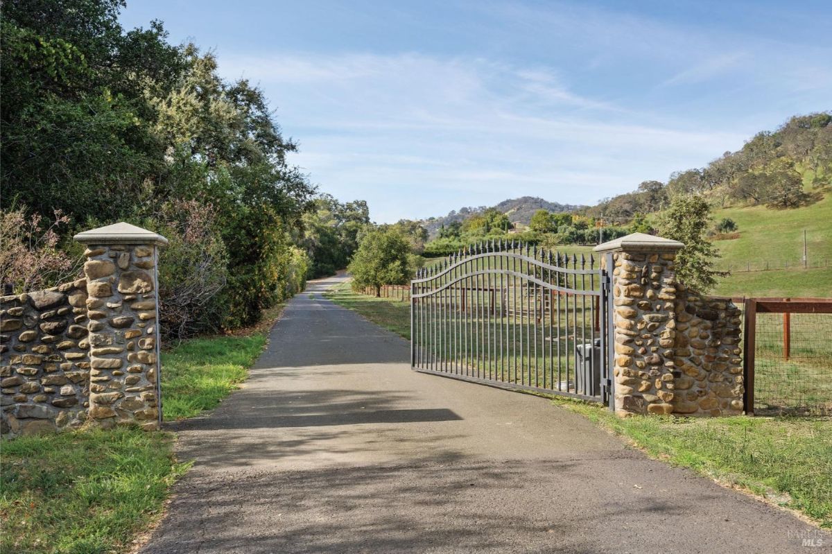 Gated entryway features wrought iron gates framed by stone pillars. Paved driveway leads through greenery and open fields toward rolling hills in the background.