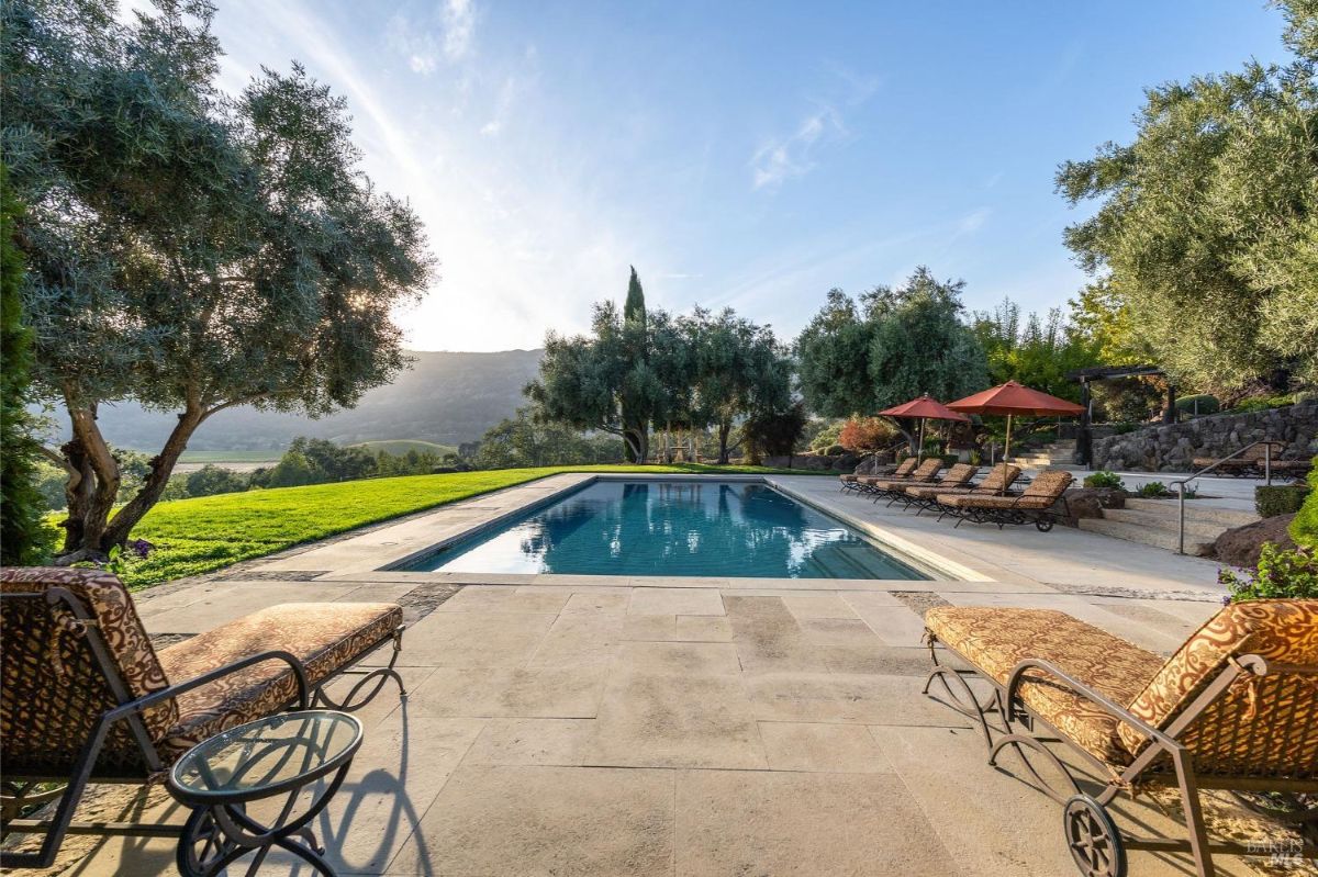 View from the edge of a pool overlooking a manicured lawn and a forested landscape, with rolling hills in the distance. The sky displays a gradient of warm and cool tones during sunset, creating a serene atmosphere.