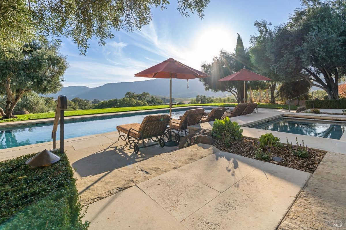 Outdoor pool area with lounge chairs and umbrellas overlooking a scenic mountain landscape. Surrounding trees and greenery frame the poolside space.