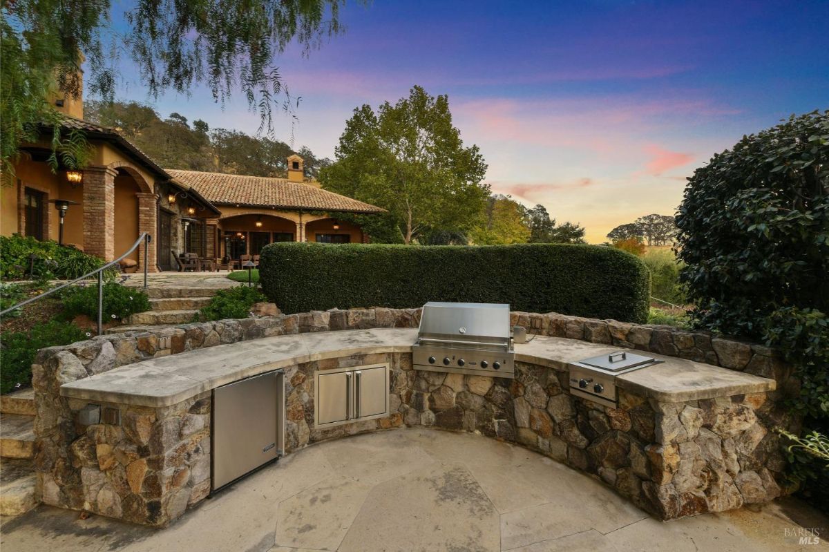 Built-in outdoor stone barbecue with a grill and appliances, set in a landscaped garden near a covered patio. The house and garden steps are visible in the background. 