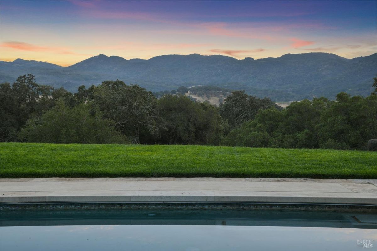 Rectangular swimming pool surrounded by a stone deck and lounge chairs, with red umbrellas providing shade. Set against a backdrop of manicured lawns, olive trees, and rolling hills, offering a serene and picturesque view.