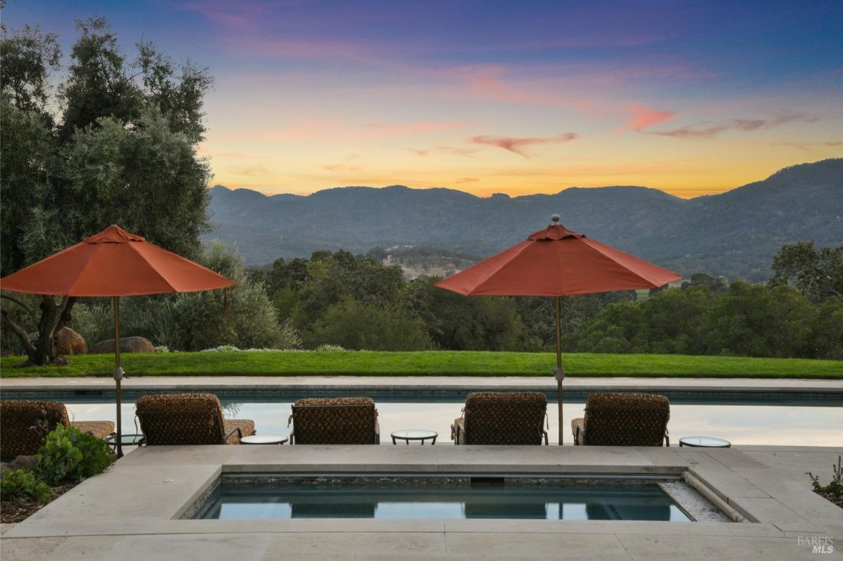 Outdoor seating area with lounge chairs and red umbrellas overlooking a rectangular pool and hot tub. The backdrop features a manicured lawn, rolling hills, and a colorful sunset sky.