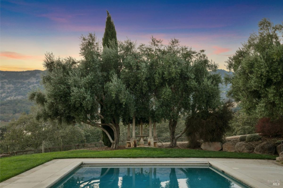 Poolside view with olive trees and a cypress tree framing a small pergola in the background. The scene captures a tranquil atmosphere at sunset, with soft lighting and distant rolling hills.