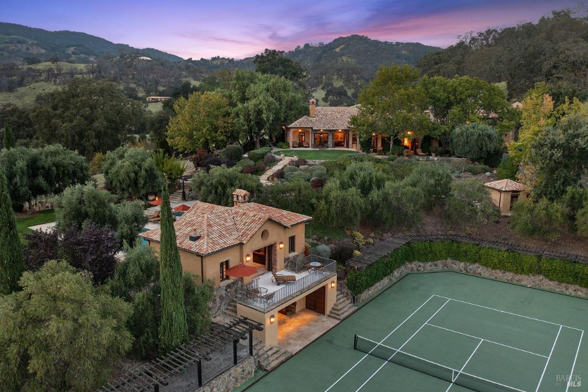 Aerial view of a villa complex with a tennis court, landscaped gardens, and a pool against a backdrop of hills.