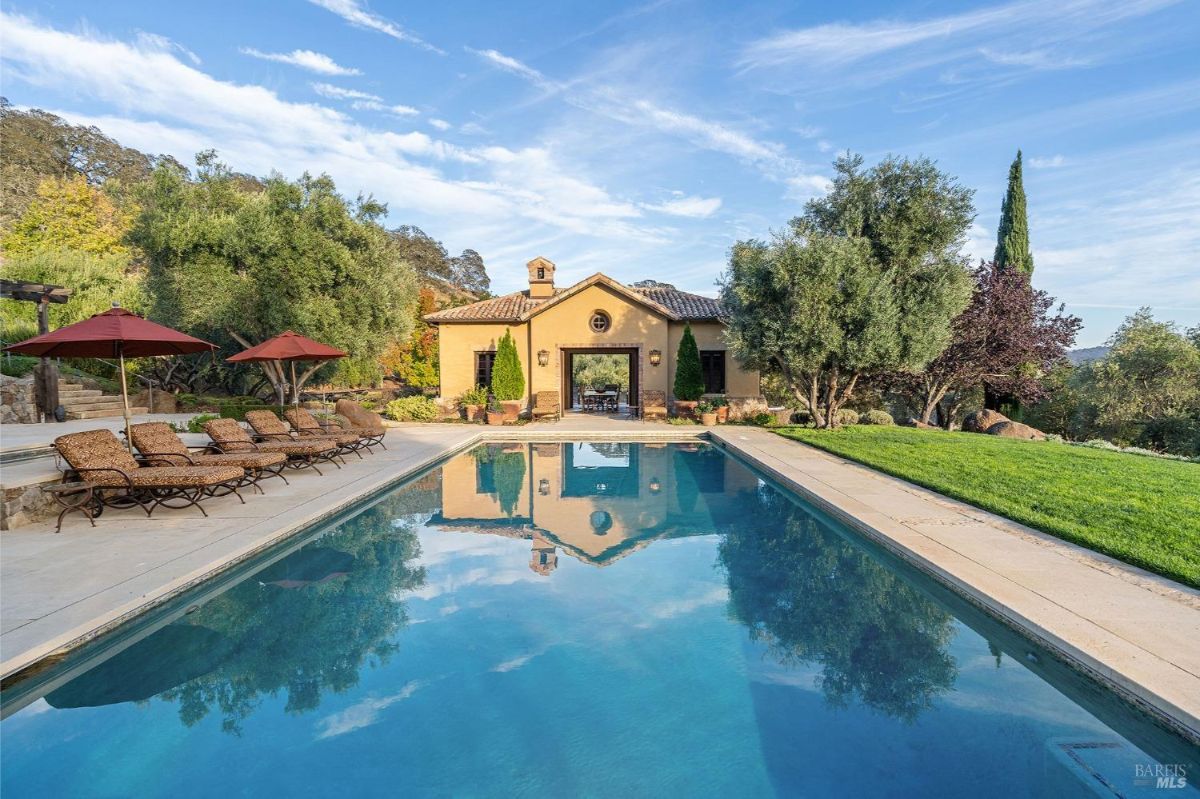 Outdoor area featuring a rectangular swimming pool with lounge chairs and umbrellas along one side. The pool is bordered by landscaped greenery and leads to a stucco structure with a tiled roof and an open archway.