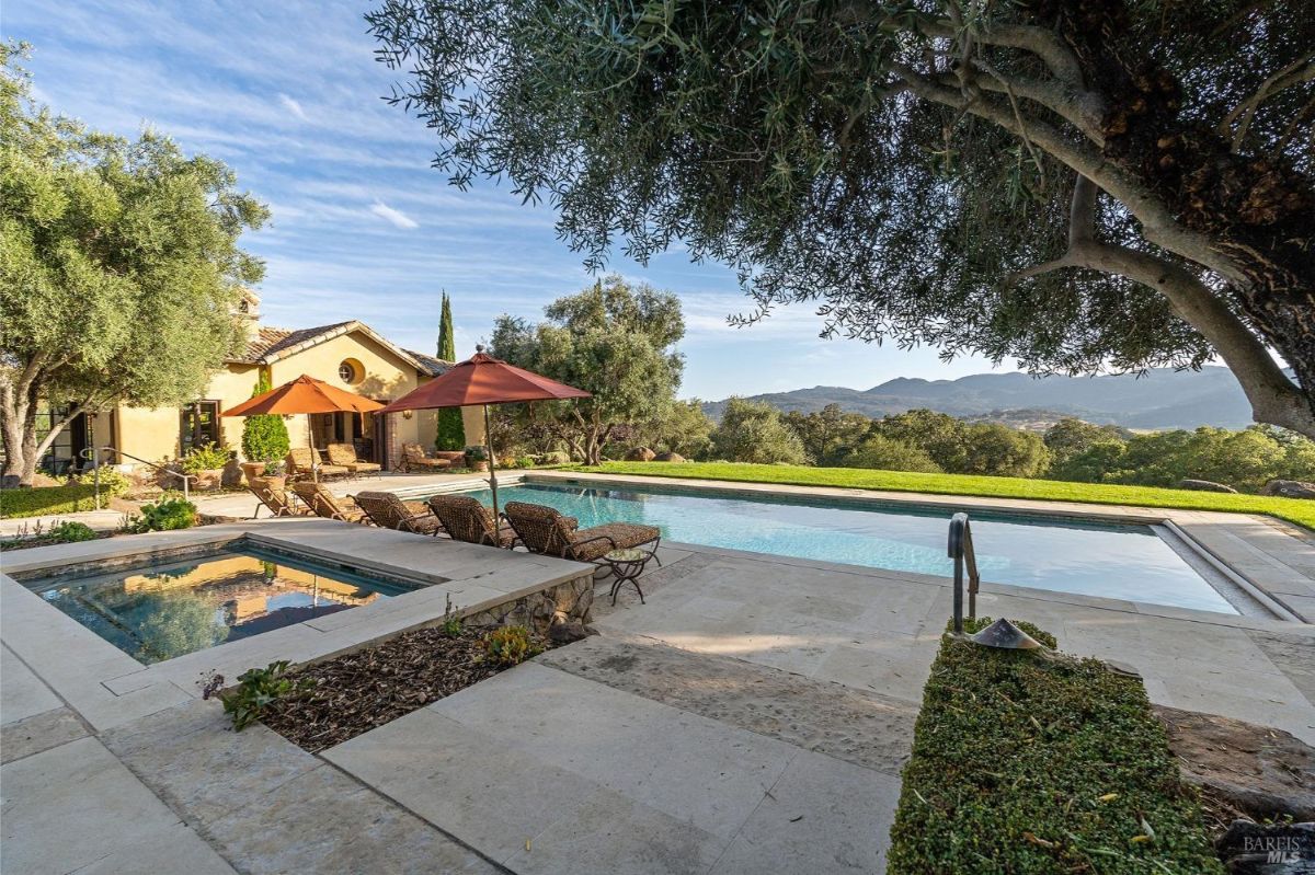 Outdoor area featuring a hot tub and a rectangular swimming pool surrounded by stone decking and lounge chairs with red umbrellas. The setting is enhanced by olive trees, manicured landscaping, and a stucco building with a tiled roof in the background.