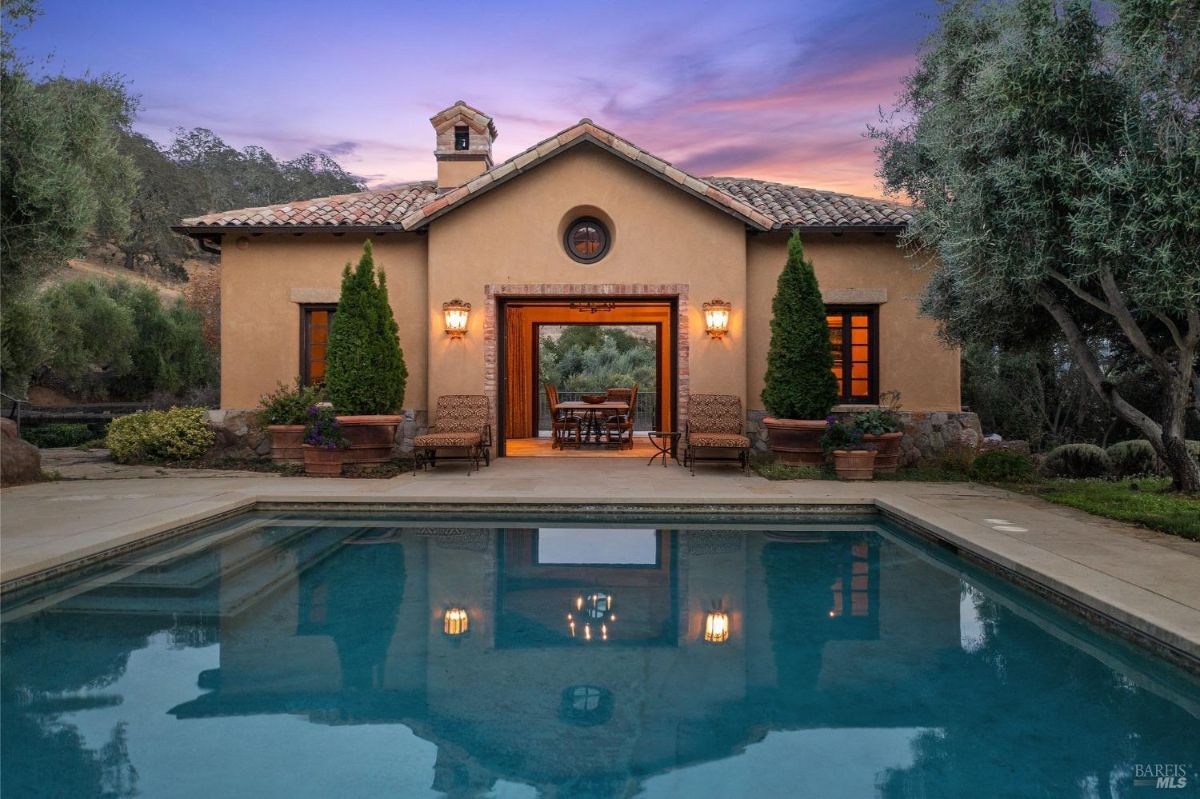 Outdoor area featuring a rectangular swimming pool with lounge chairs and umbrellas along one side. The pool is bordered by landscaped greenery and leads to a stucco structure with a tiled roof and an open archway.