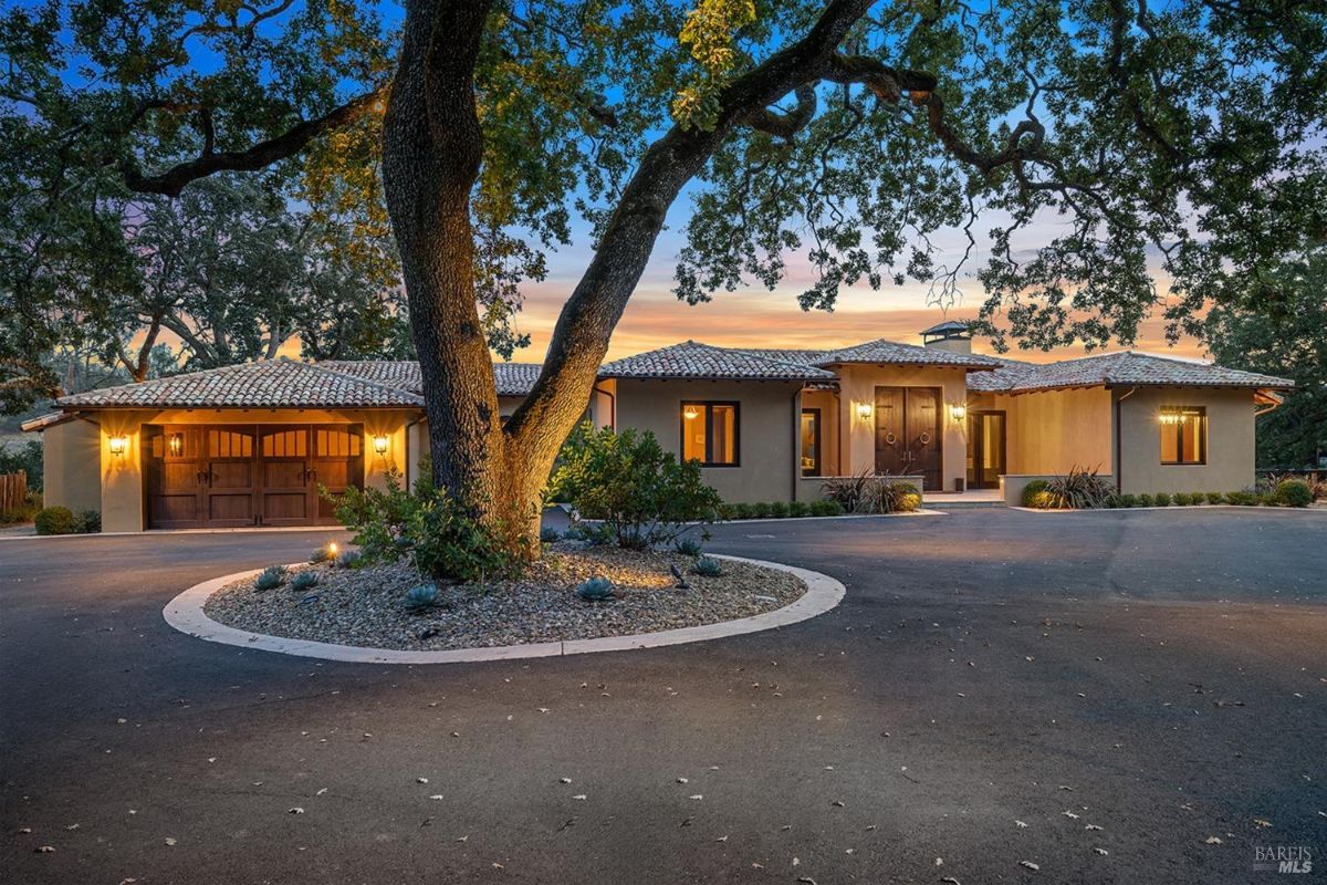 Circular driveway surrounding a large oak tree leads to a single-story home with a tiled roof.