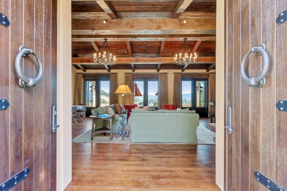 Wooden double doors open to a living room featuring exposed wooden beams and chandeliers. Large windows and doors at the far end provide natural light and views of the outdoor scenery.