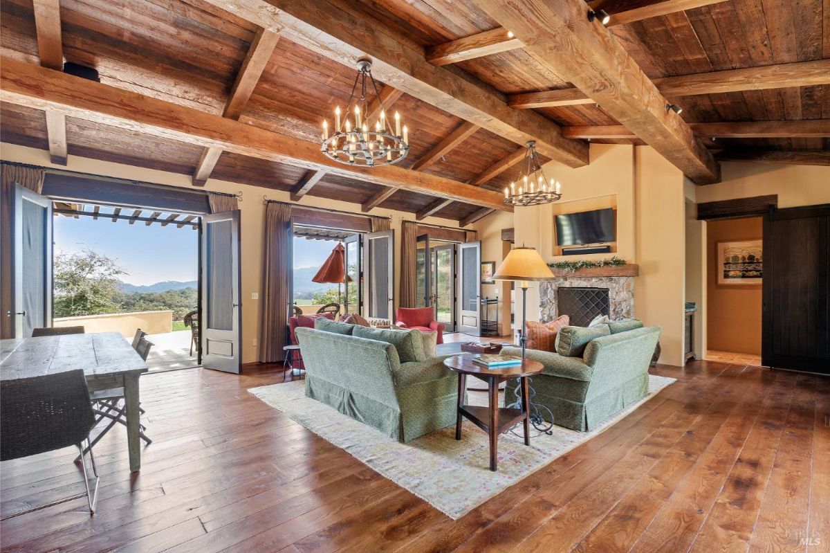 Room features exposed wooden beams, chandeliers, and hardwood flooring. Multiple doors open to an outdoor patio with mountain views, while furniture includes sofas, a table, and a stone fireplace.