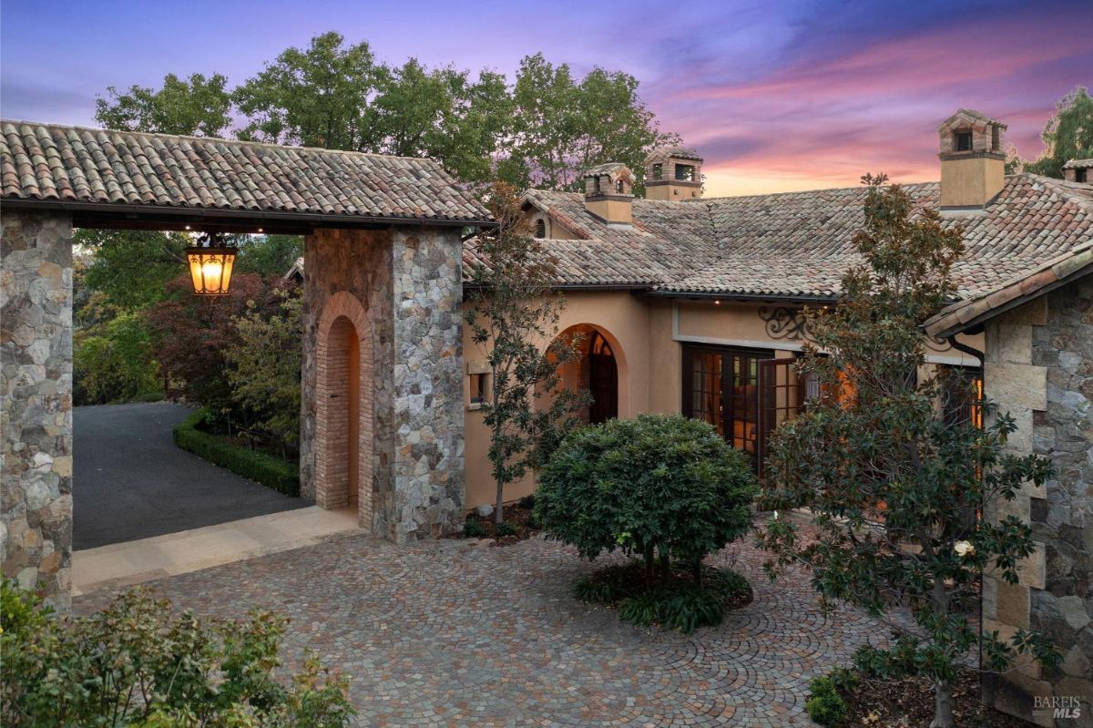 Stone entry archway leading to a cobblestone driveway and a Mediterranean-style home with tiled roofing.