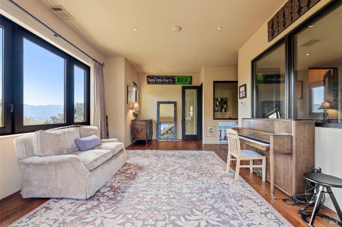 A cozy room features a wooden piano and a comfortable beige sofa placed near large windows with expansive views. The decor includes a patterned rug, a wall mirror, and framed artwork, creating an inviting atmosphere for relaxation or music.