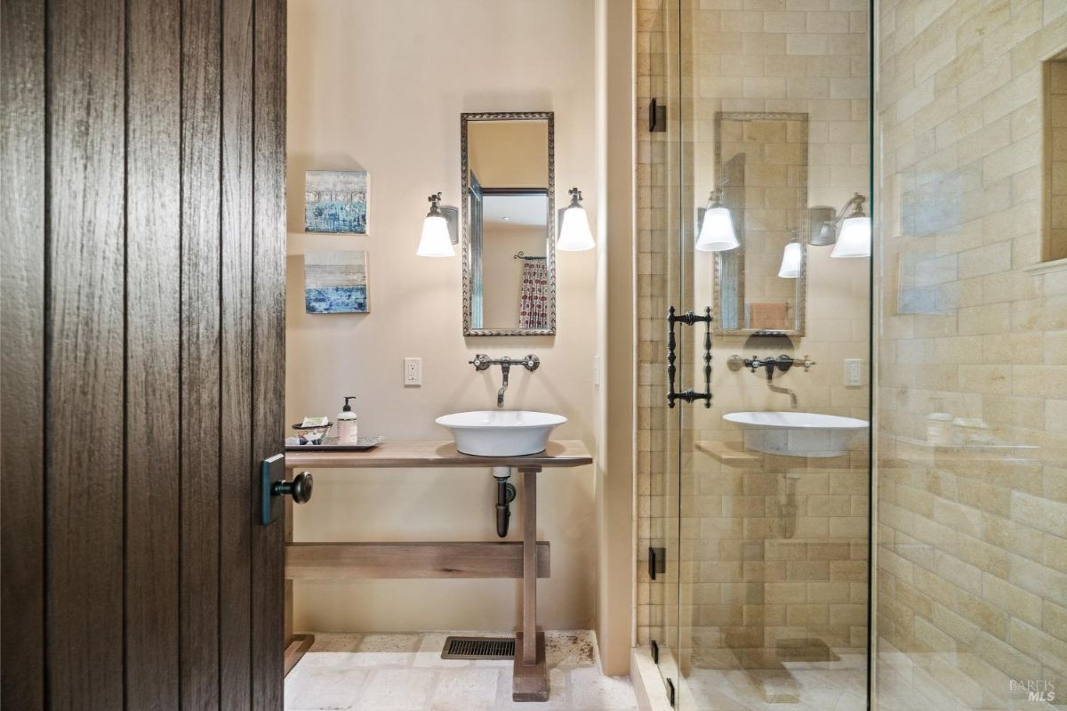 Bathroom with a walk-in glass shower and beige tiled walls. Features a floating wood vanity with a vessel sink, wall-mounted faucet, and decorative lighting fixtures.