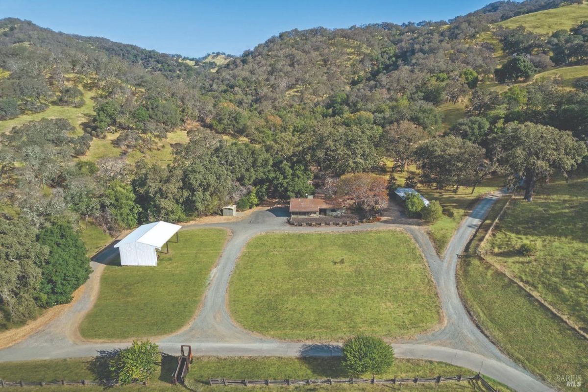 Ranch property includes a central house surrounded by trees and open fields. Features a white barn structure, gravel driveways, and fenced grassy areas.