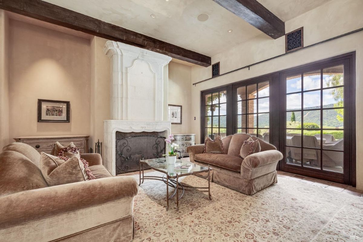 Living room features a white carved fireplace and large glass doors opening to an outdoor patio. Furnishings include plush sofas, a patterned rug, and a glass coffee table with metal framing.