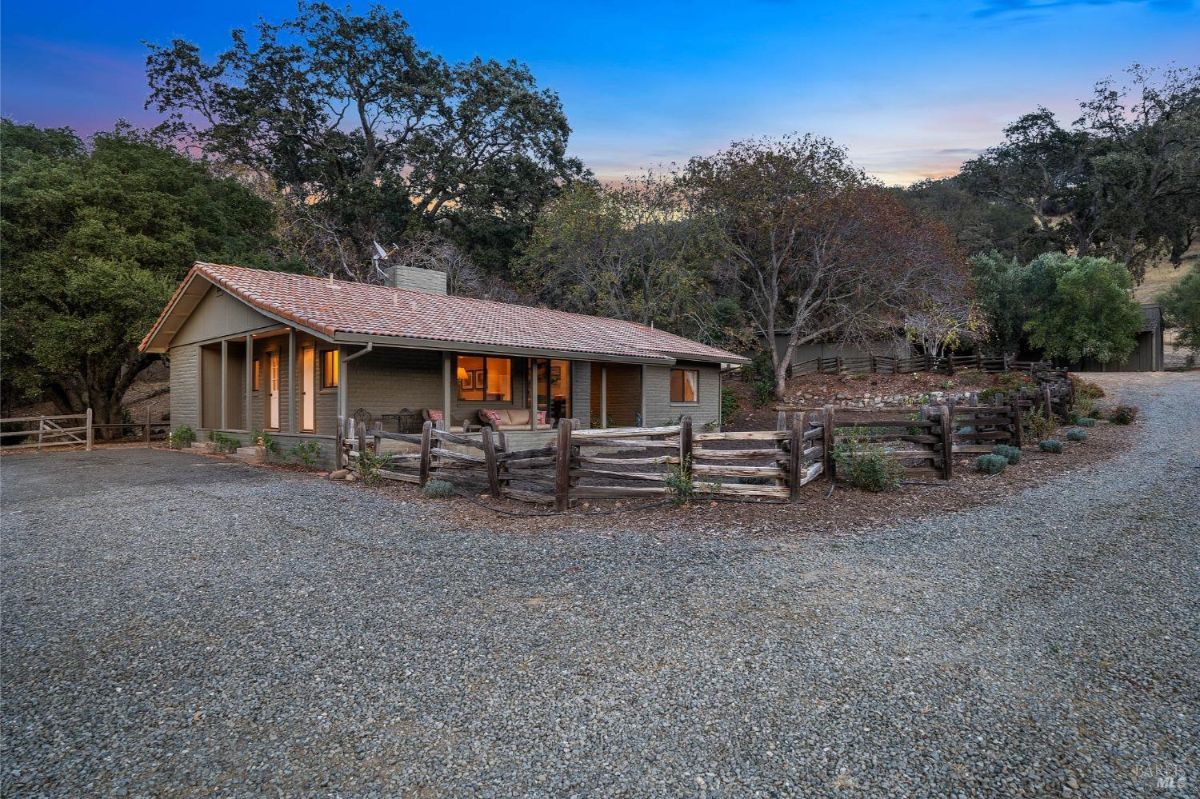 Ranch-style home with a gravel driveway and a wooden fenced area in a rural setting.