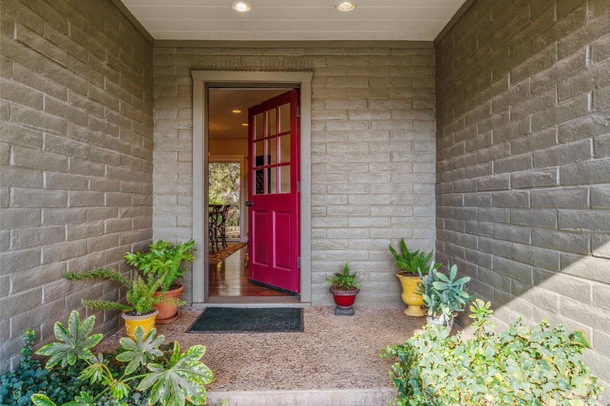 Entryway features a red door with glass panes, opening into a room with visible hardwood floors. Brick walls surround the space, and potted plants add greenery to the exterior.