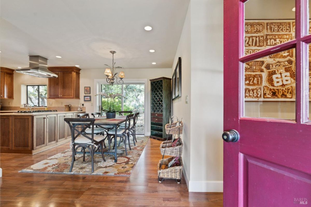 Dining area with hardwood flooring features a rectangular table with black chairs and a floral rug. Kitchen with wooden cabinets, a stainless steel range hood, and access to a backyard through glass doors is visible in the background.