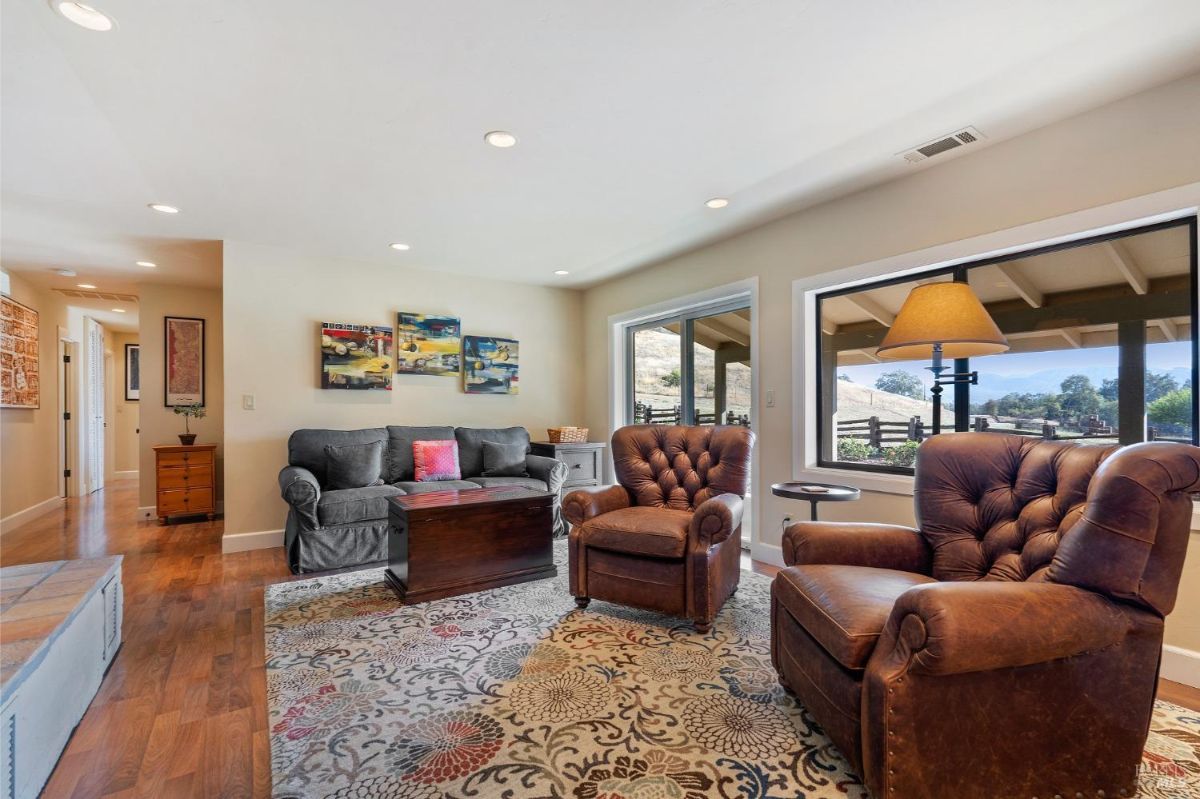 Living area features a gray upholstered sofa, two leather armchairs, and a patterned area rug. Large windows and glass doors provide natural light and views of the outdoor landscape.