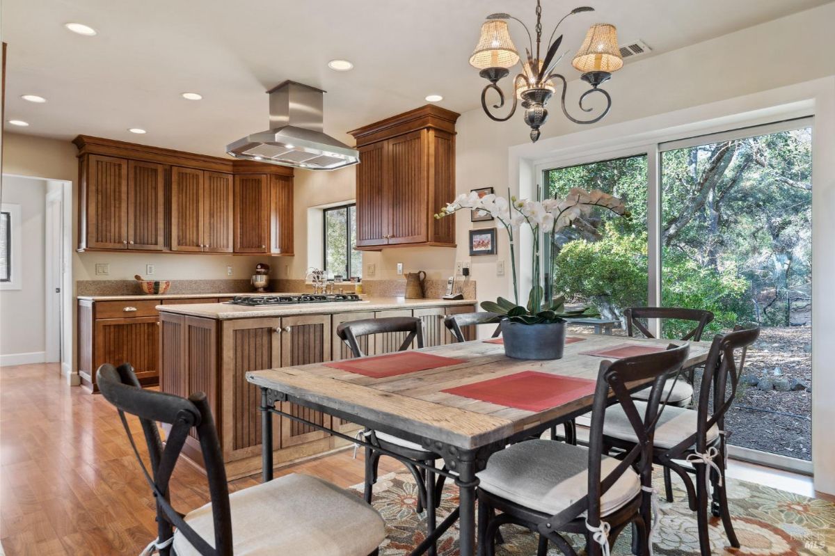 A spacious kitchen and dining area features wooden cabinets, a center island with a cooktop, and a rustic dining table with metal-framed chairs. Large sliding glass doors offer views of greenery and natural light fills the room.