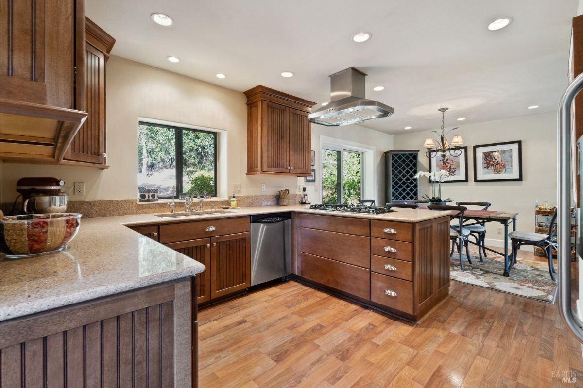 A kitchen layout features wood cabinetry, granite countertops, and a central island with a built-in gas cooktop. Adjacent dining space includes large windows and a sliding glass door that offers views of the outdoor surroundings.