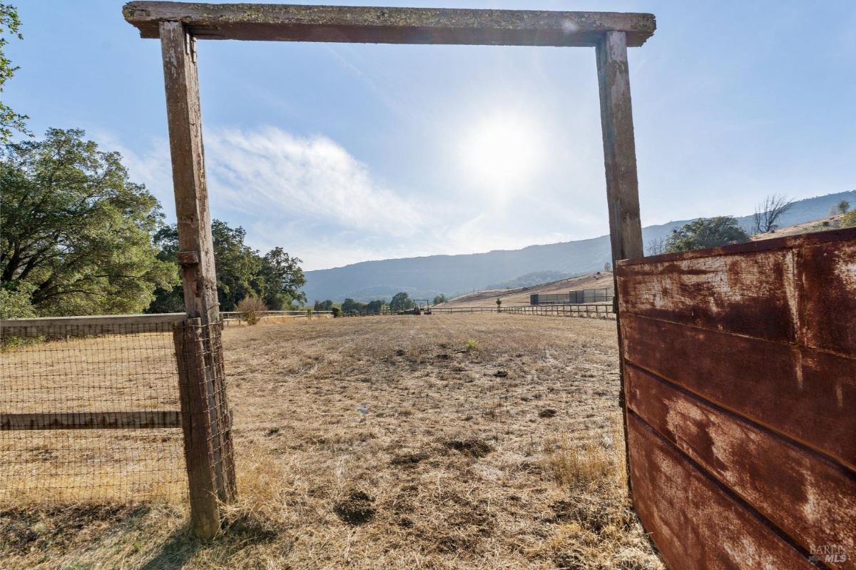 A rustic wooden gate leading to a vast, open field. The field is dry and brown, with a chain-link fence running along its edge. Beyond the field, rolling hills stretch into the distance, and a clear blue sky with fluffy white clouds completes the scene. 