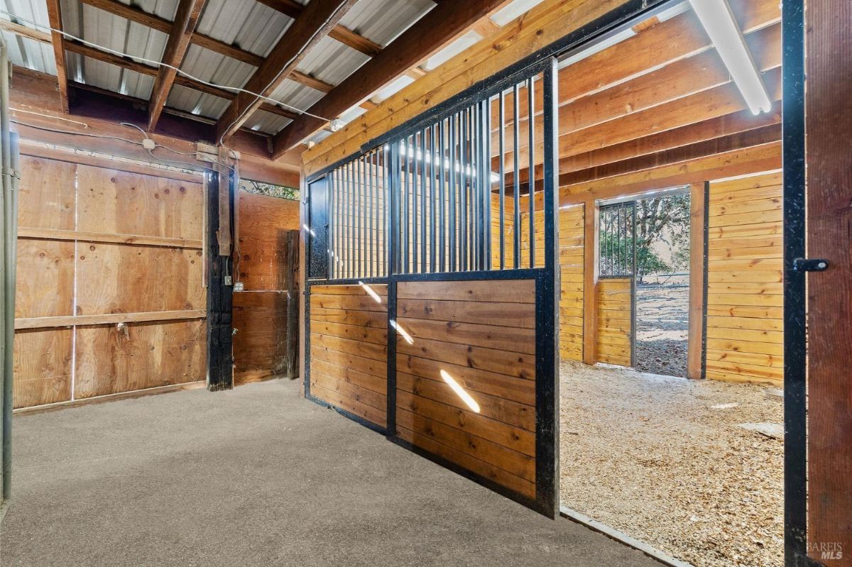 A wooden and metal horse stable with multiple stalls, lit by natural light through open doors.