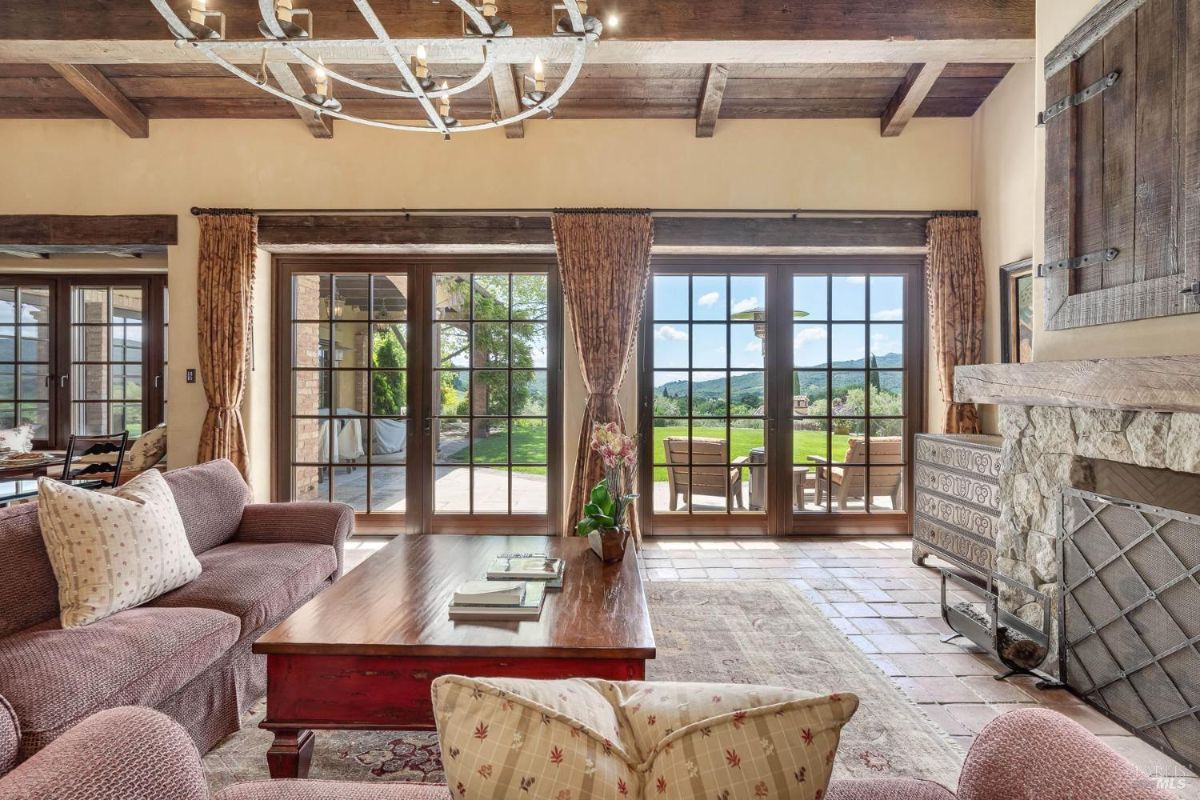 Living room features large glass doors providing views of the patio and surrounding landscape. Furnishings include a wooden coffee table, patterned upholstery, and exposed wooden beams on the ceiling.