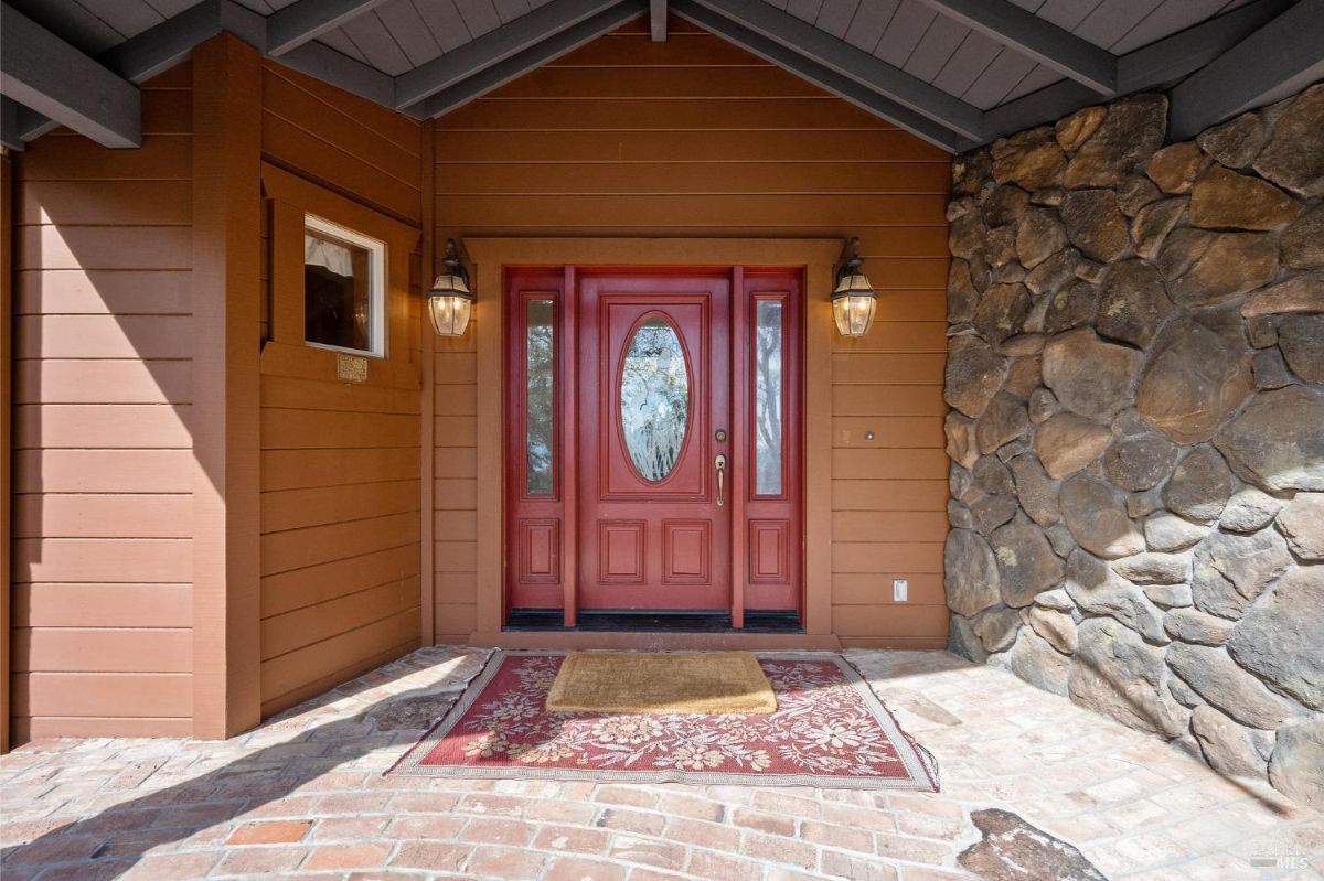 Covered entryway features a red door with decorative glass panels and brick flooring. Stone and wood siding create a rustic facade with wall-mounted lantern lights.