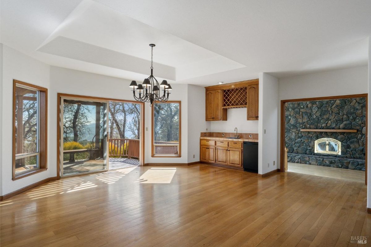 An open dining and living space features hardwood floors, a stone fireplace, and built-in wooden cabinetry with a small sink and wine rack. Large windows and a sliding glass door lead to an outdoor deck and offer natural light and wooded views.