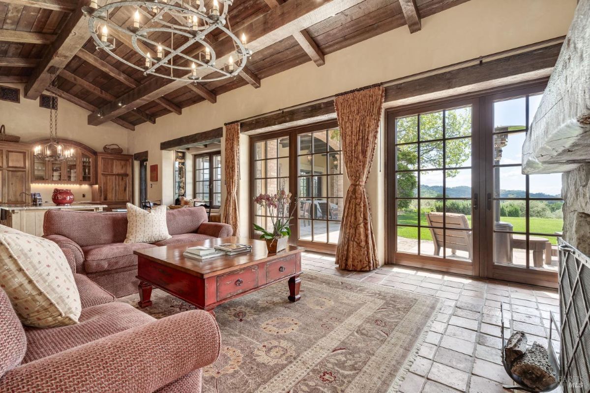 Living area features exposed wood beam ceilings and large glass doors opening to an outdoor patio. Furnishings include pink upholstered chairs, a red coffee table, and a patterned rug, with a view of greenery outside.