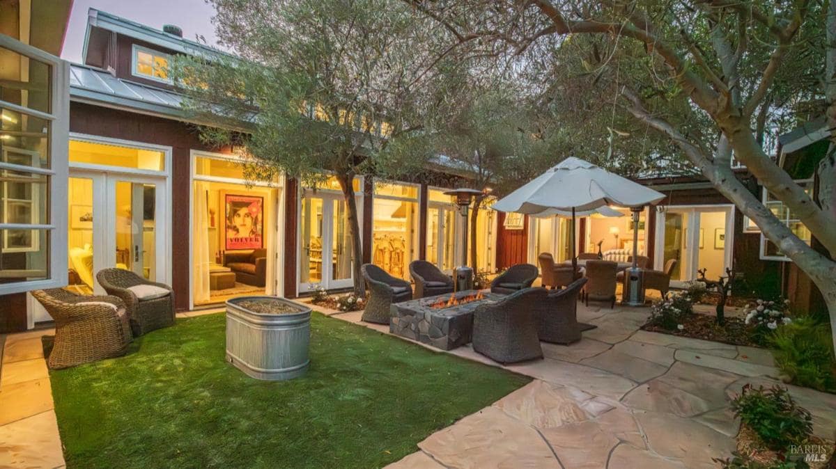 Outdoor seating area with fire pit surrounded by wicker chairs and trees, adjacent to a building with large windows.