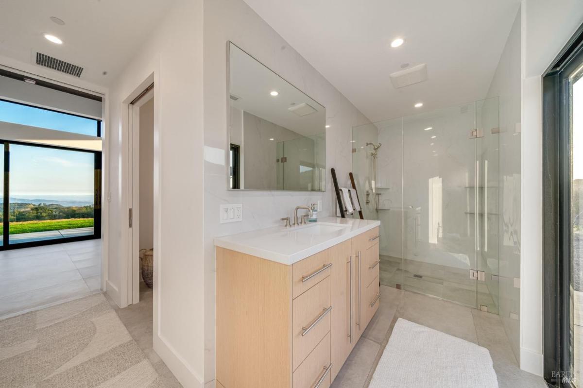 A bathroom with a double vanity and glass-enclosed shower, adjacent to a bedroom with outdoor views.