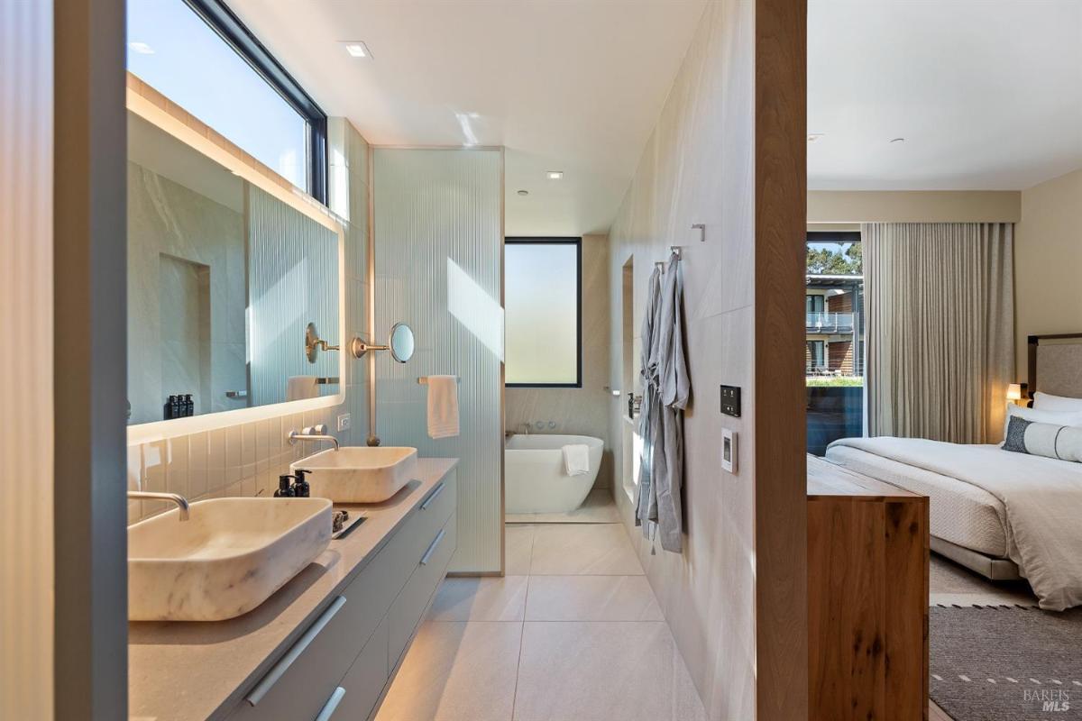 A bathroom with double sinks, a freestanding bathtub, and a view into the bedroom.