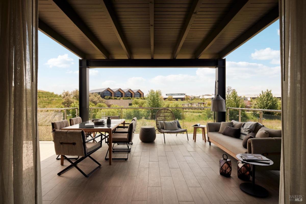 A covered outdoor space with a dining table, seating area, and glass railing overlooking greenery.