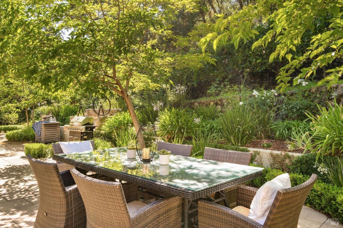 An outdoor dining with dining table with chairs is set in a garden surrounded by plants and trees.