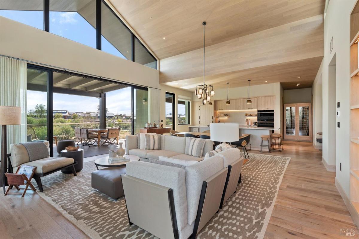 A living area with a high ceiling, large windows, and an open layout connected to a kitchen.