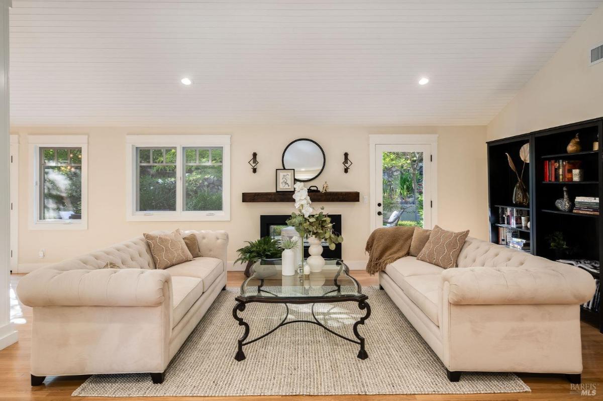 A living room features two sofas facing a coffee table, with a fireplace and windows providing natural light.

