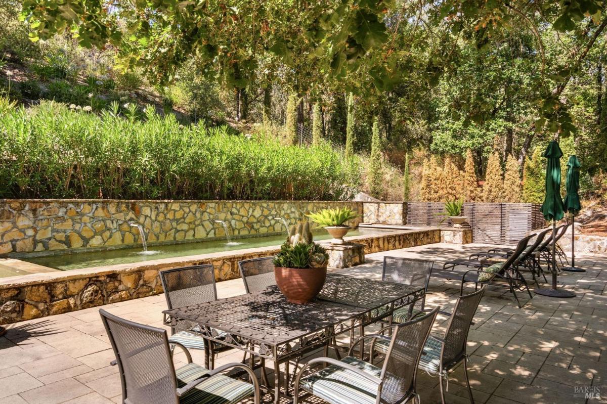 A patio area with a stone fountain, dining table, and lounge chairs surrounded by greenery.

