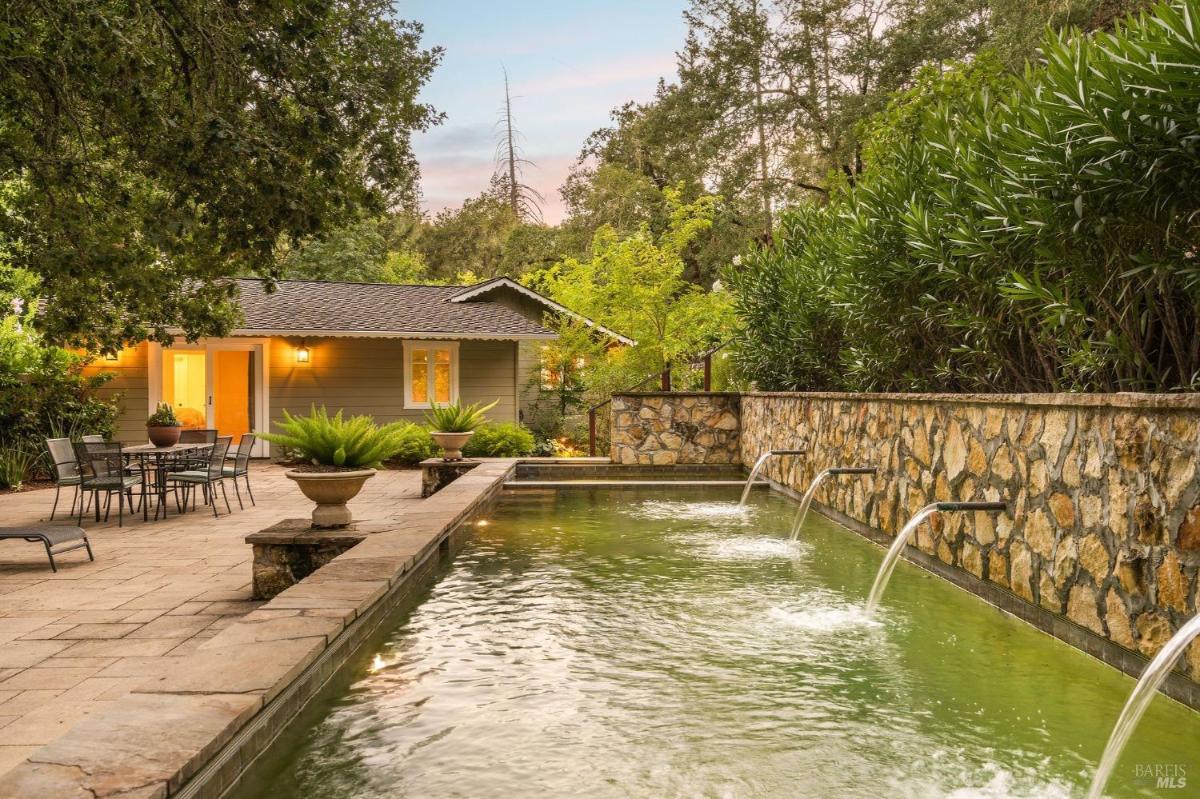 A stone patio with a pool featuring water fountains, surrounded by greenery and a house in the background.

