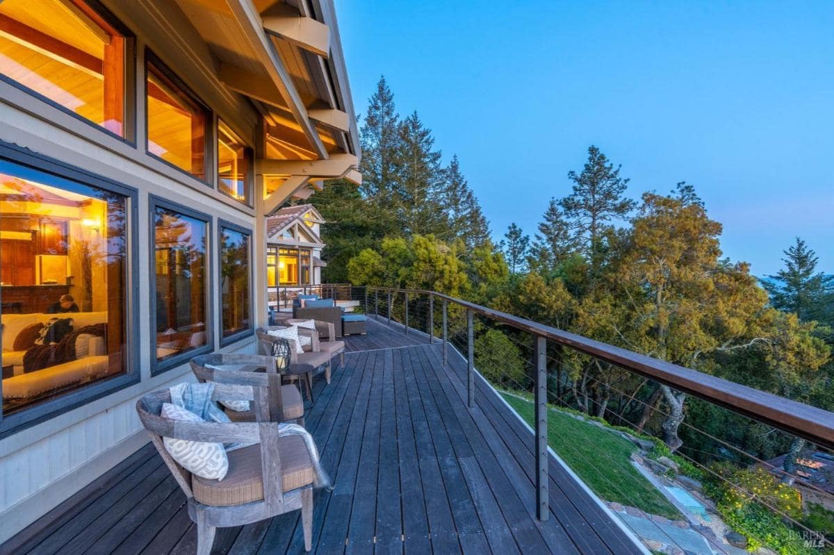 A wooden deck with chairs and a railing overlooking a forested area.