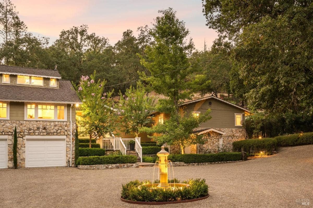 A driveway with a central fountain, leading to a house with a stone facade and surrounding greenery.

