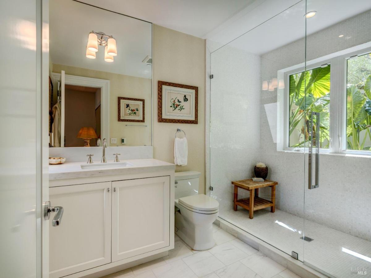 A bathroom with a large glass shower, white vanity, and a small wooden stool.