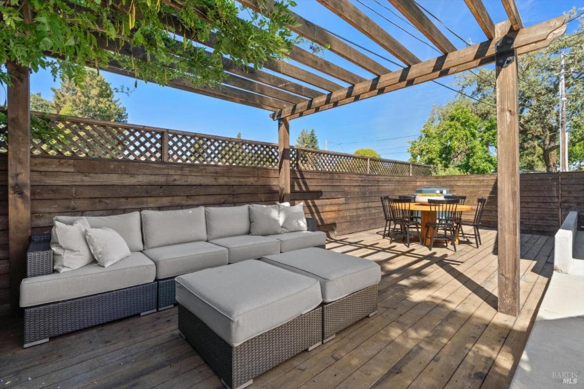 Outdoor seating area with a pergola, sofa, and a dining table on a wooden deck surrounded by a privacy fence.