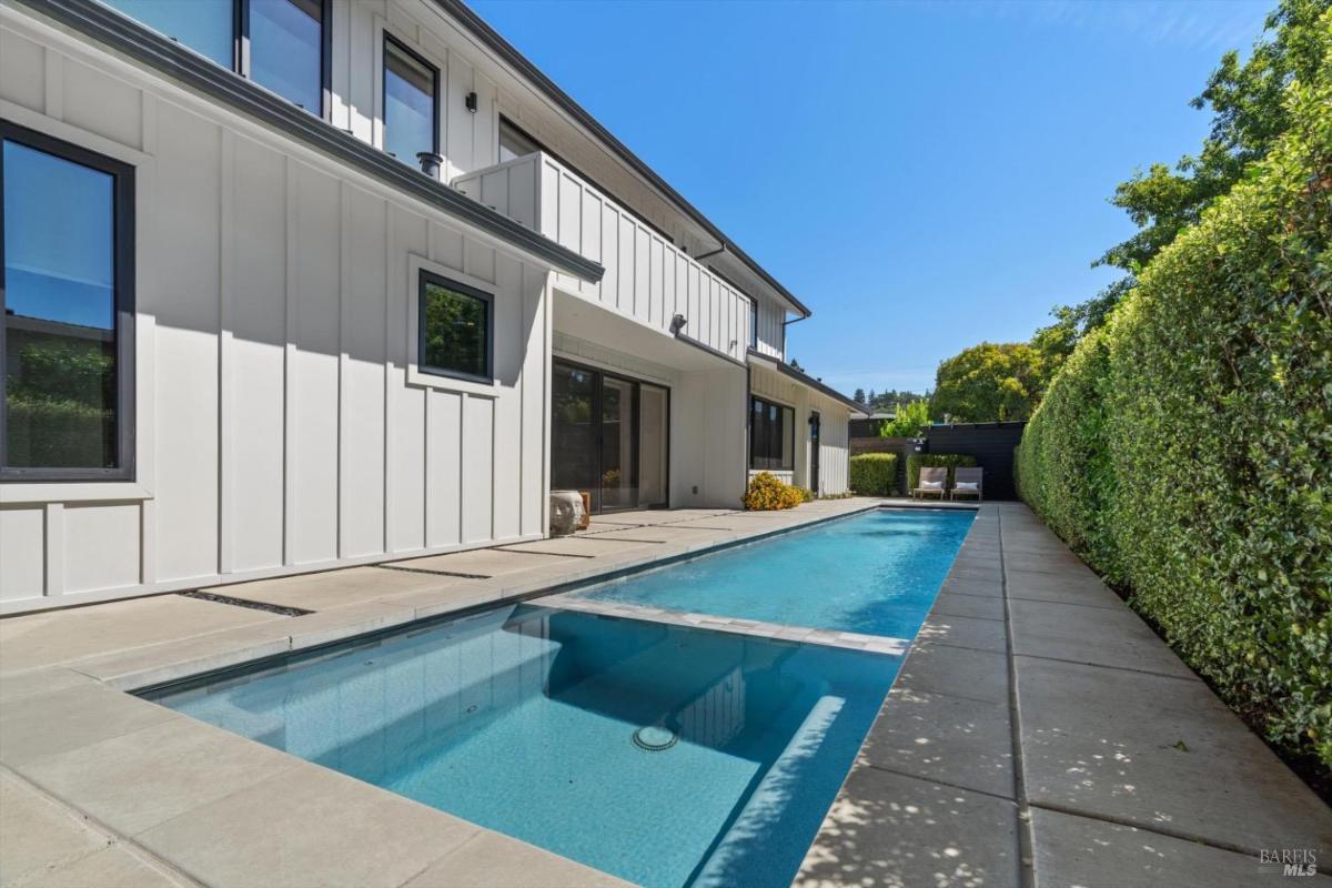 Long swimming pool with a concrete patio alongside a white house and trimmed hedges.