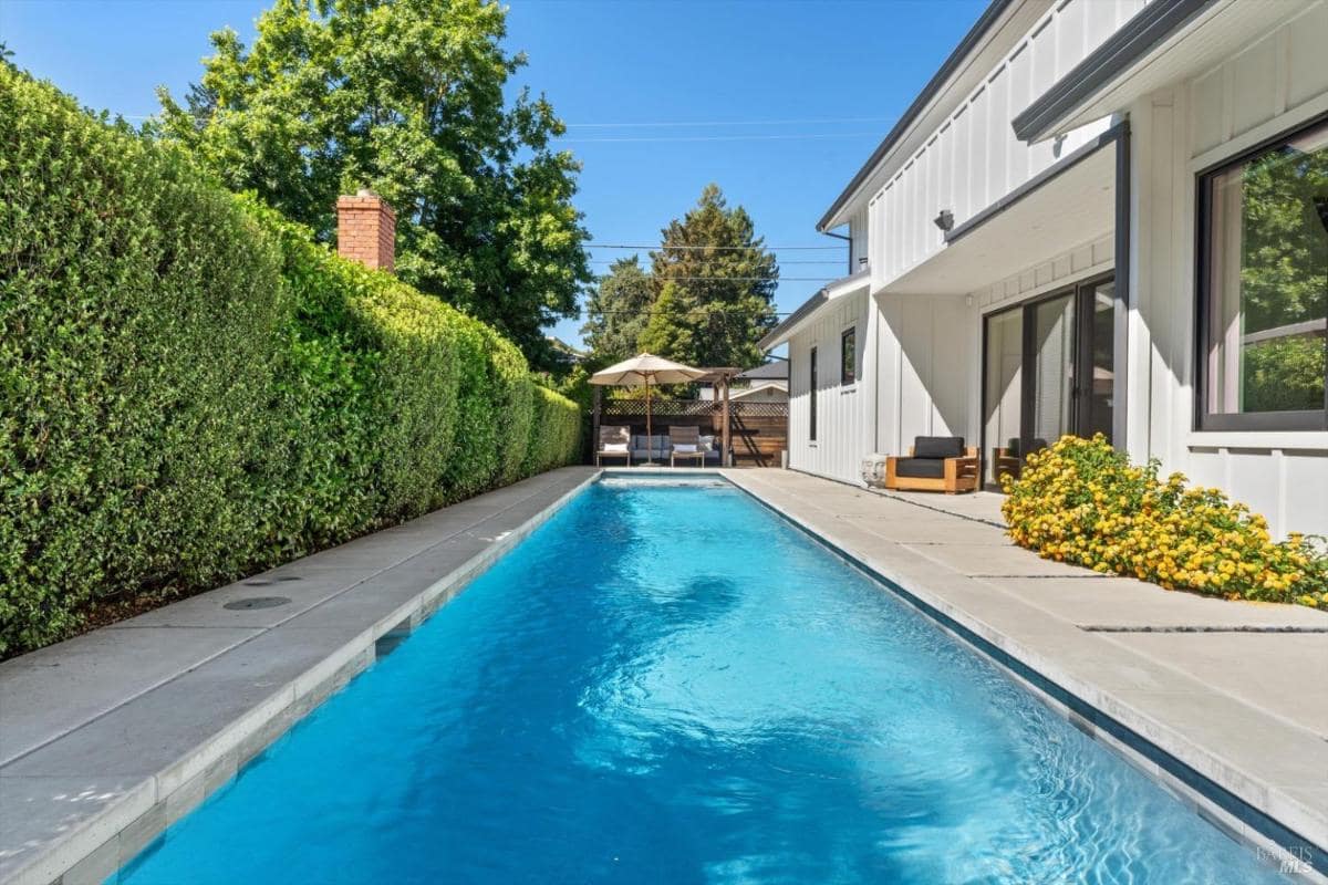 Long swimming pool with a concrete patio alongside a white house and trimmed hedges.