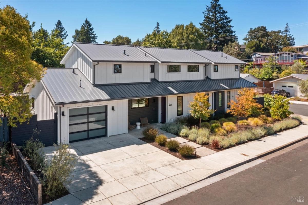 Corner view of a modern house with metal roof, white siding, and landscaped yard with shrubs.